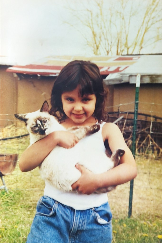 Tricia is smiling and holding her cat Birdie. There's a fence and shed in the background.
